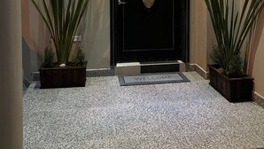 Una puerta negra con un ventanal ovalado decorativo, flanqueada por dos macetas a cada lado, que da acceso a una zona de entrada con suelo de baldosas y un felpudo.