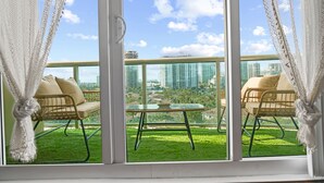 A balcony with two wicker chairs and a glass table, overlooking a cityscape and a grassy area.
