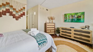 A neatly made bed with a patterned throw, a television displaying a waterfall, and a wooden dresser with a plant.