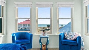 Ein Zimmer mit drei großen Fenstern mit Blick auf einen Strand, zwei blaue Sessel, ein kleiner Tisch mit einem Korb und die Ansicht eines Gebäudes mit rotem Dach.