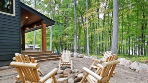 Une terrasse en bois, dotée de chaises Adirondack et d’un foyer, entourée d’une zone boisée.