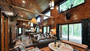 L’intérieur d’un chalet rustique, avec un plafond en bois, un canapé en cuir, une table basse ronde et une grande fenêtre donnant sur une forêt.