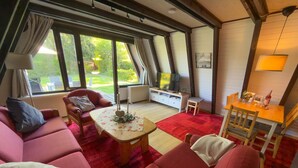Un salon chaleureux avec un canapé rouge, une table basse en bois et un téléviseur posé sur un meuble blanc. De grandes fenêtres offrent une vue sur un jardin.