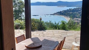 A covered outdoor seating area with a table and chairs, overlooking a coastal landscape with a view of the sea and distant hills.