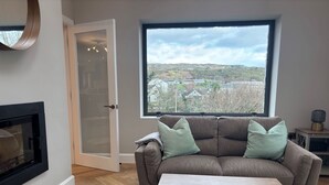 Ein Wohnzimmer mit Ecksofa, Couchtisch, Kamin und einem großen Fenster mit Blick auf eine hügelige Landschaft.