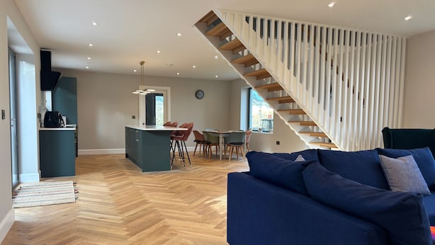 Ein moderner Wohnraum mit Fischgrätparkett, einem blauen Sofa, einer Kücheninsel und einer Treppe mit weißen Geländern.