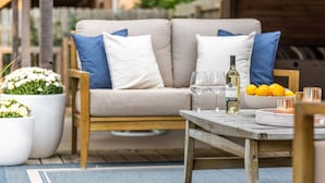 Ein gemütlicher Außenbereich mit einem Holztisch, einem beigefarbenen Sofa mit blauen Kissen und einem Blumentopf.