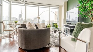 Una sala de estar moderna con un sofá gris, un sillón blanco y una mesa de centro de vidrio. Un ventanal ofrece vistas a un horizonte urbano.