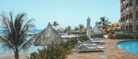 Zwembadgedeelte van een resort met ligbedden, palmbomen en een gebouw met balkons.