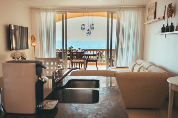 Un salon moderne avec un canapé, une table basse et un coin cuisine équipé d’un évier et d’une machine à café. Un balcon offre une vue sur la mer.