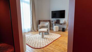 A hotel room with a wooden floor, a round table, a chair, a TV mounted on the wall, and a window with curtains.