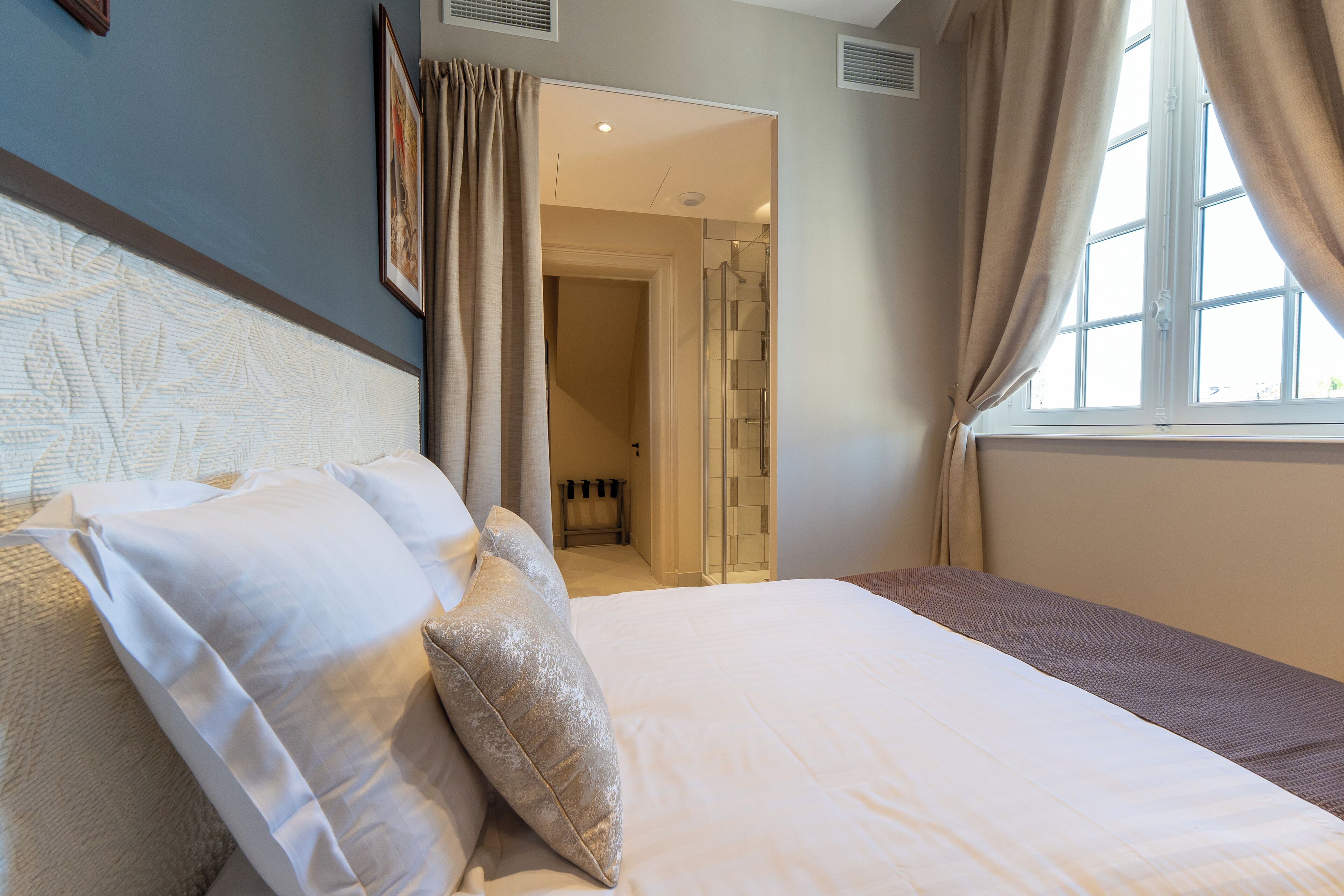 Una cama bien hecha con ropa de cama blanca y color beige, un cuadro en la pared y una ventana con cortinas.