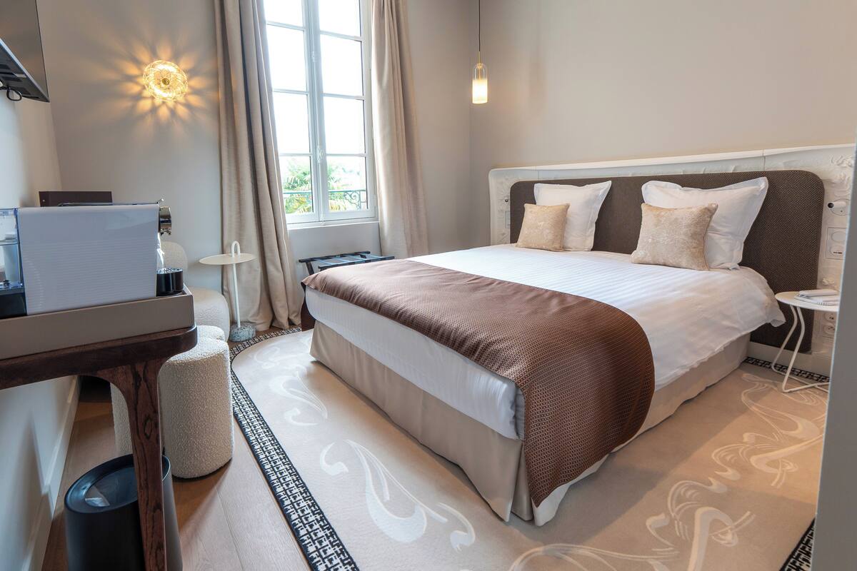 A hotel room with a large bed, a desk with a computer, a small table, and a trash bin.