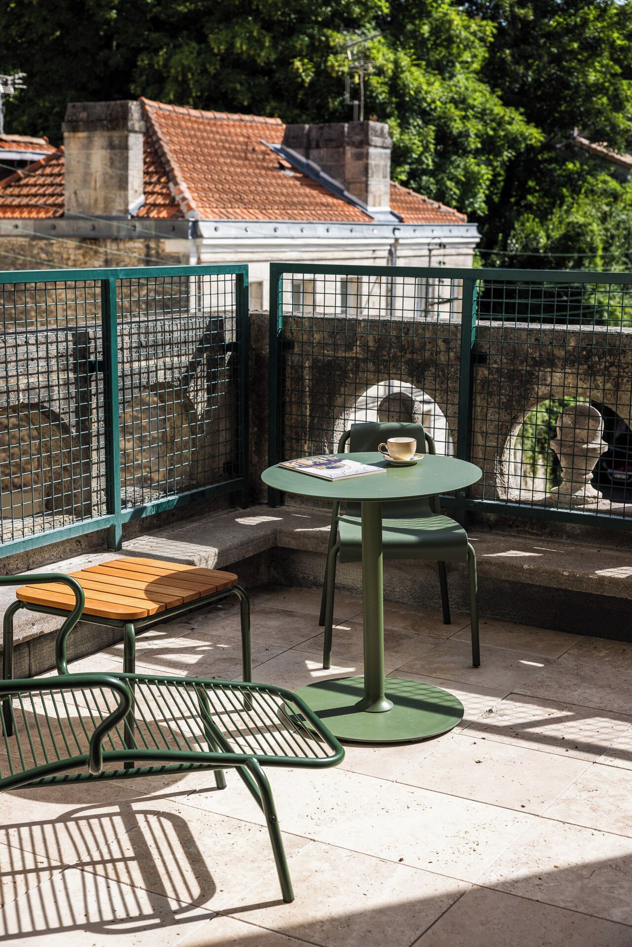 Una mesa y sillas de metal verde en un espacio al aire libre, con un muro de piedra y un techo de tejas al fondo.