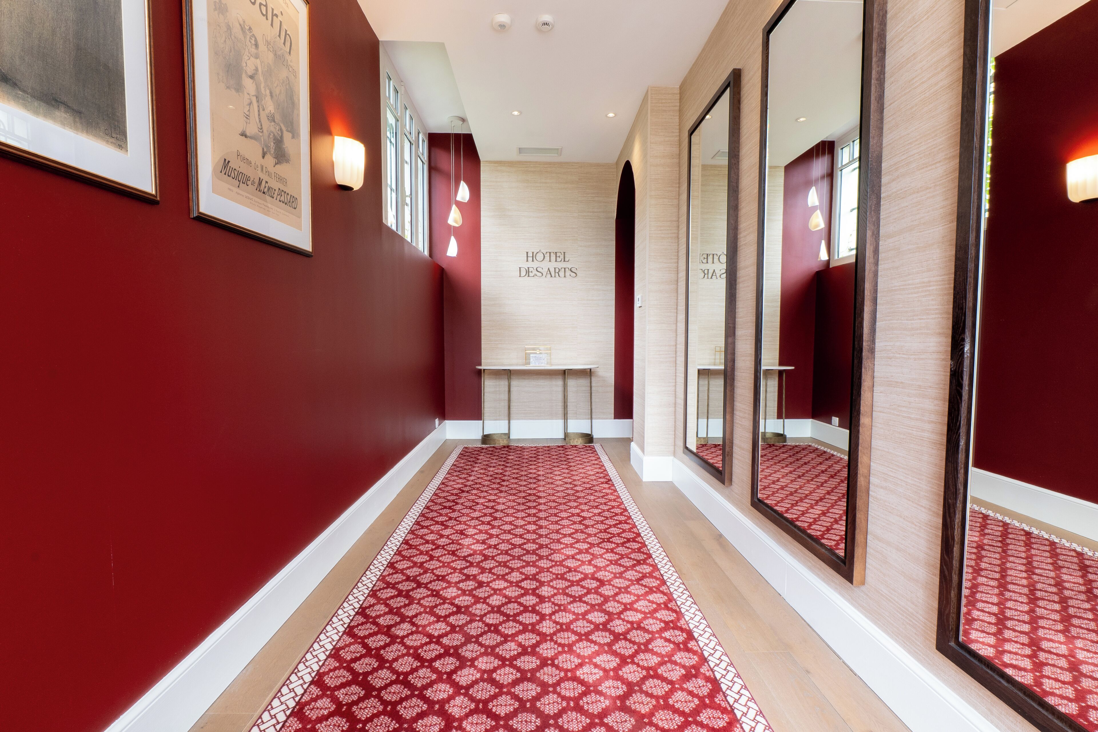 Un couloir avec une moquette rouge, des œuvres d’art encadrées accrochées aux murs et une enseigne indiquant « HÔTEL DES ARTS ».