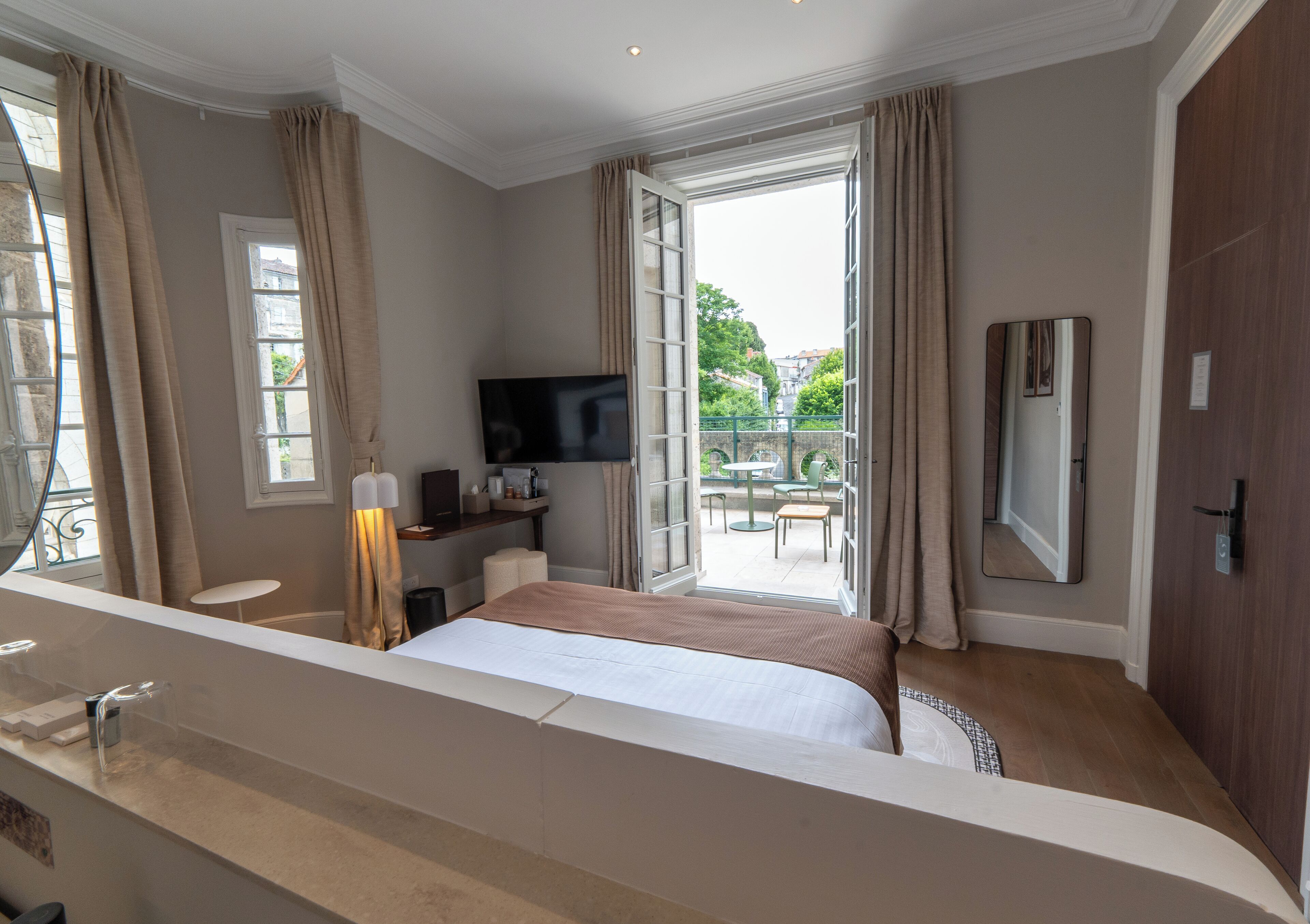 Quarto de hotel com cama, televisão, escrivaninha e varanda com mesa e cadeiras.