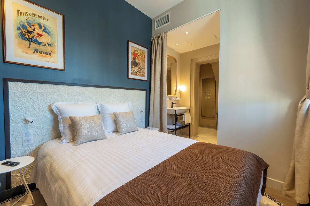A hotel room with a large bed, a bedside table, a nightstand, a bathroom with a sink and mirror, and a framed poster on the wall.