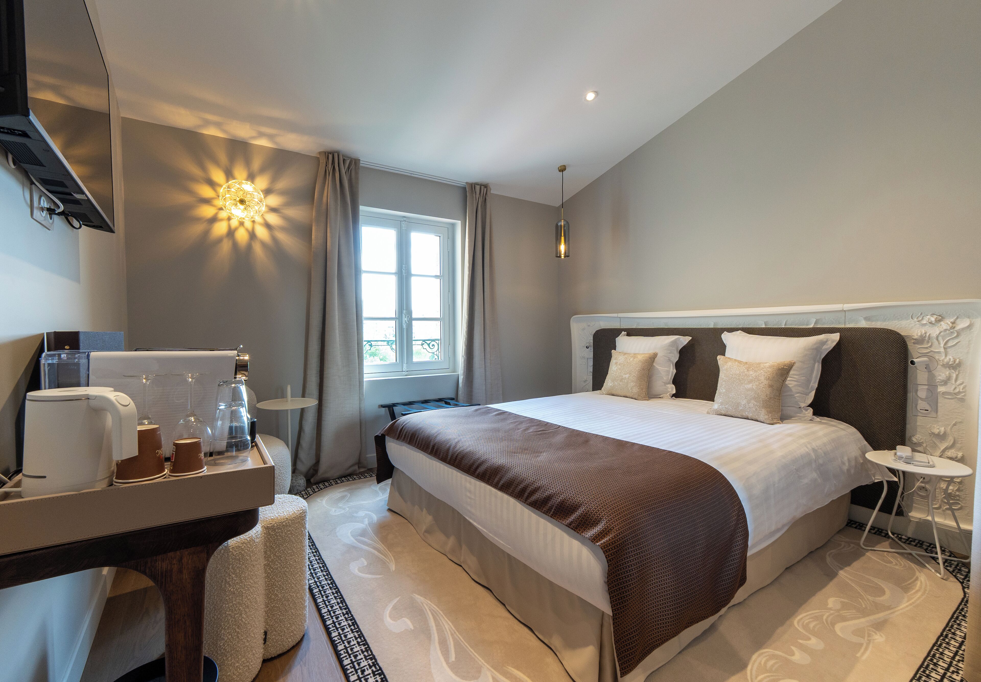 A hotel room with a large bed, a bedside table, a desk with a coffee maker, and a window with curtains.