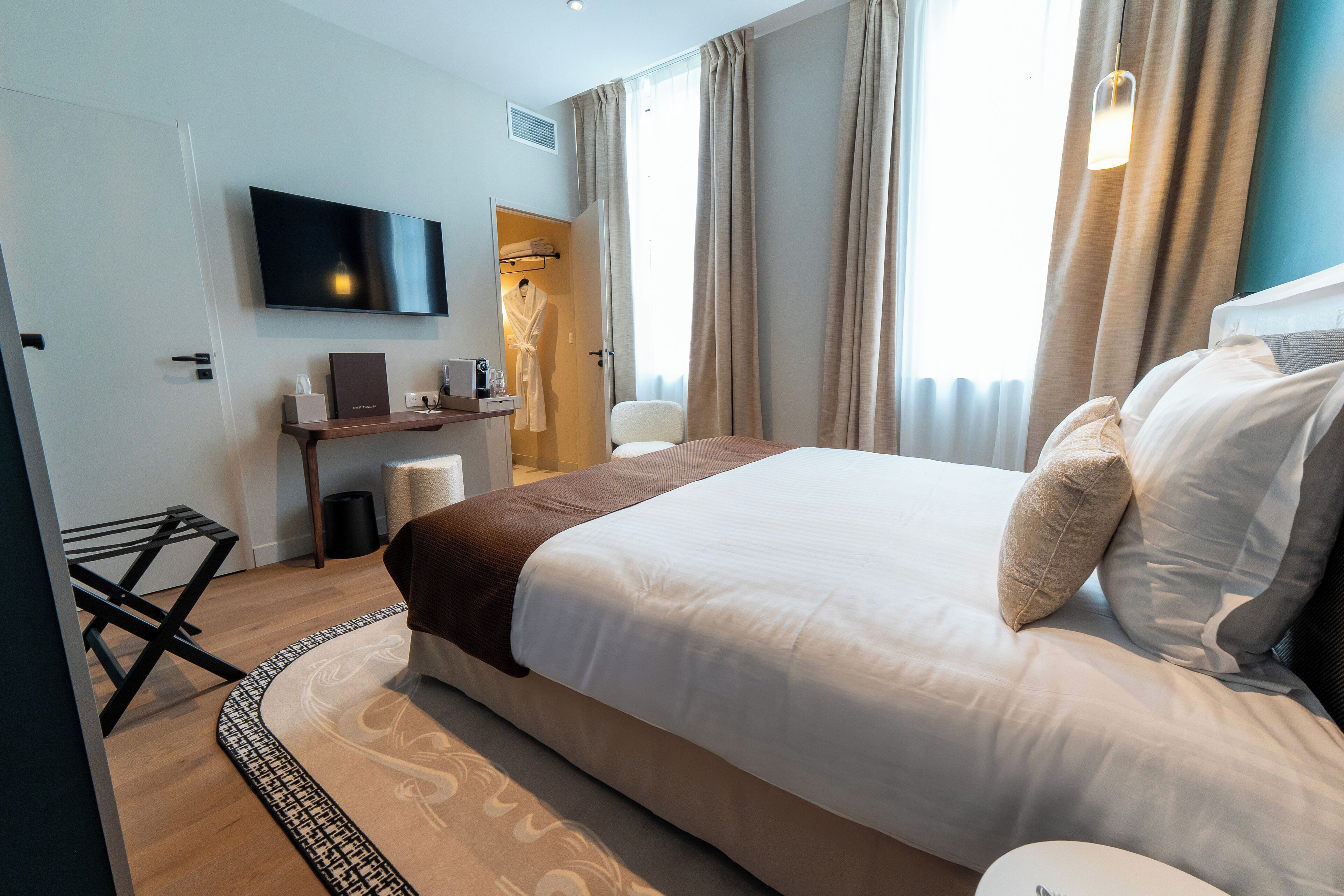 A hotel room with a large bed, a TV mounted on the wall, a desk with a chair, and a window with curtains.
