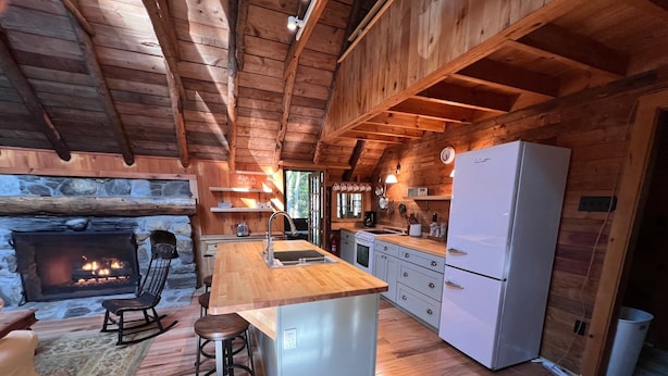 A cozy cabin interior with a stone fireplace, wooden floors, a kitchen with a refrigerator, and a dining area with a rocking chair. - Two charming cottages, a quiet ocean cove, a serene forest with private trails (Phippsburg)