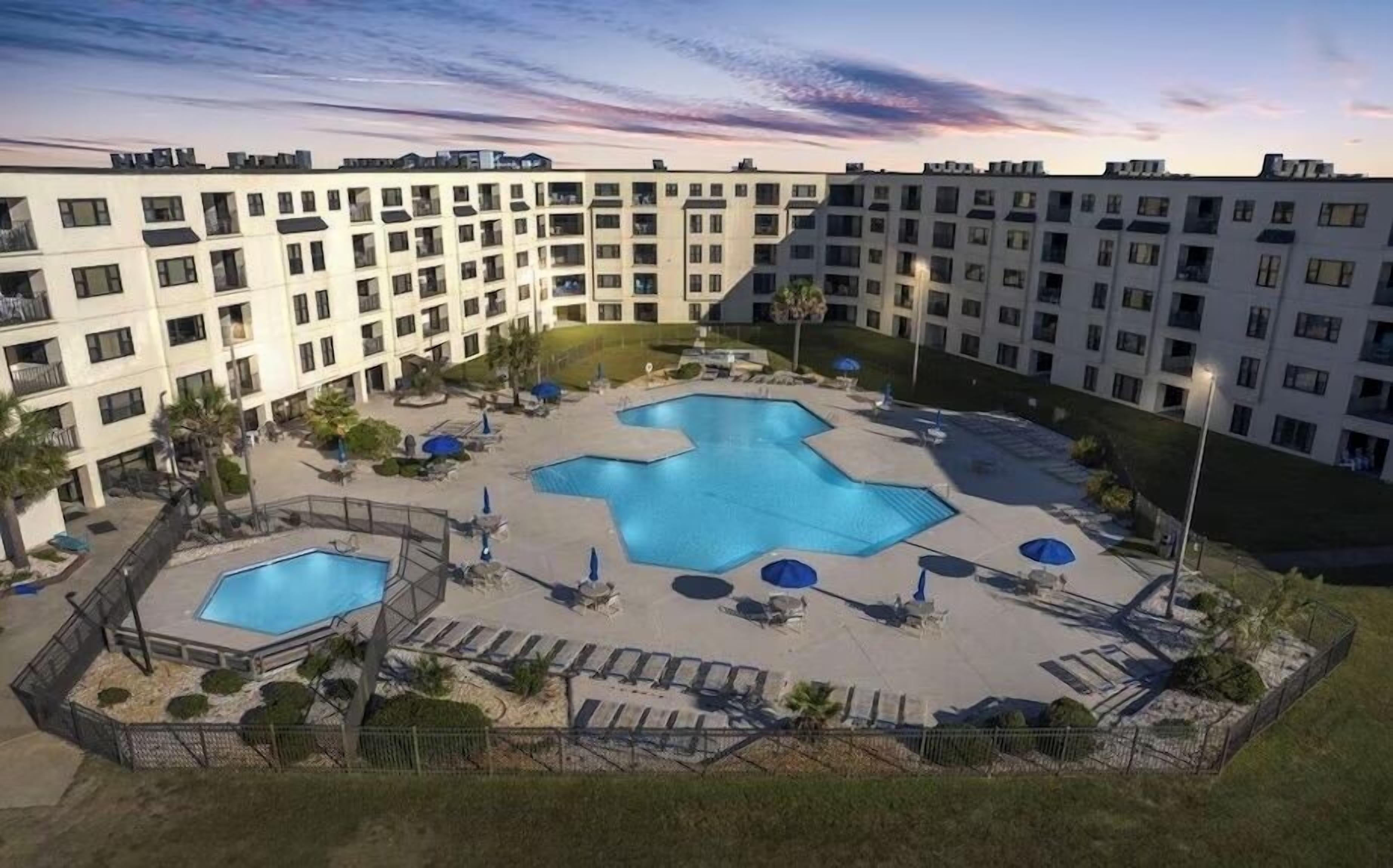 A multi-story hotel with a central courtyard featuring two swimming pools, surrounded by lounge chairs and umbrellas.