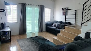 Ein geräumiges Wohnzimmer mit einer großen schwarzen Couch, einer kleineren Couch, einem Couchtisch und einem Fernseher auf einem Schrank. Eine Treppe mit Geländer führt zu einer oberen Etage.