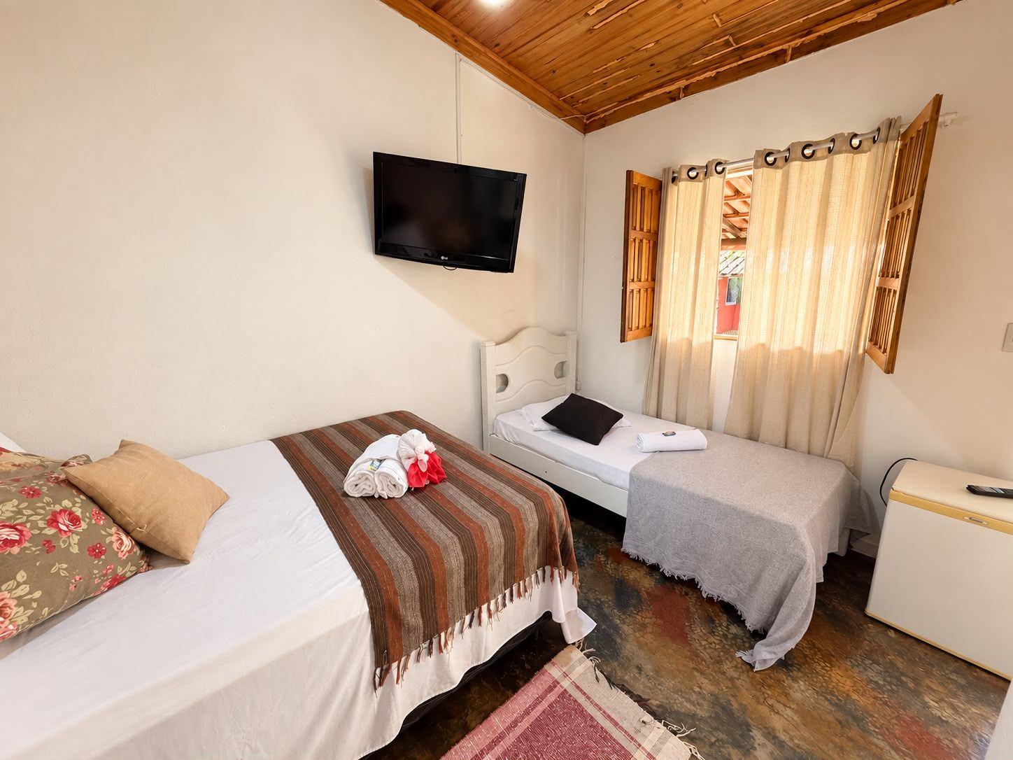 A bedroom with two beds, a television mounted on the wall, and a window with curtains.