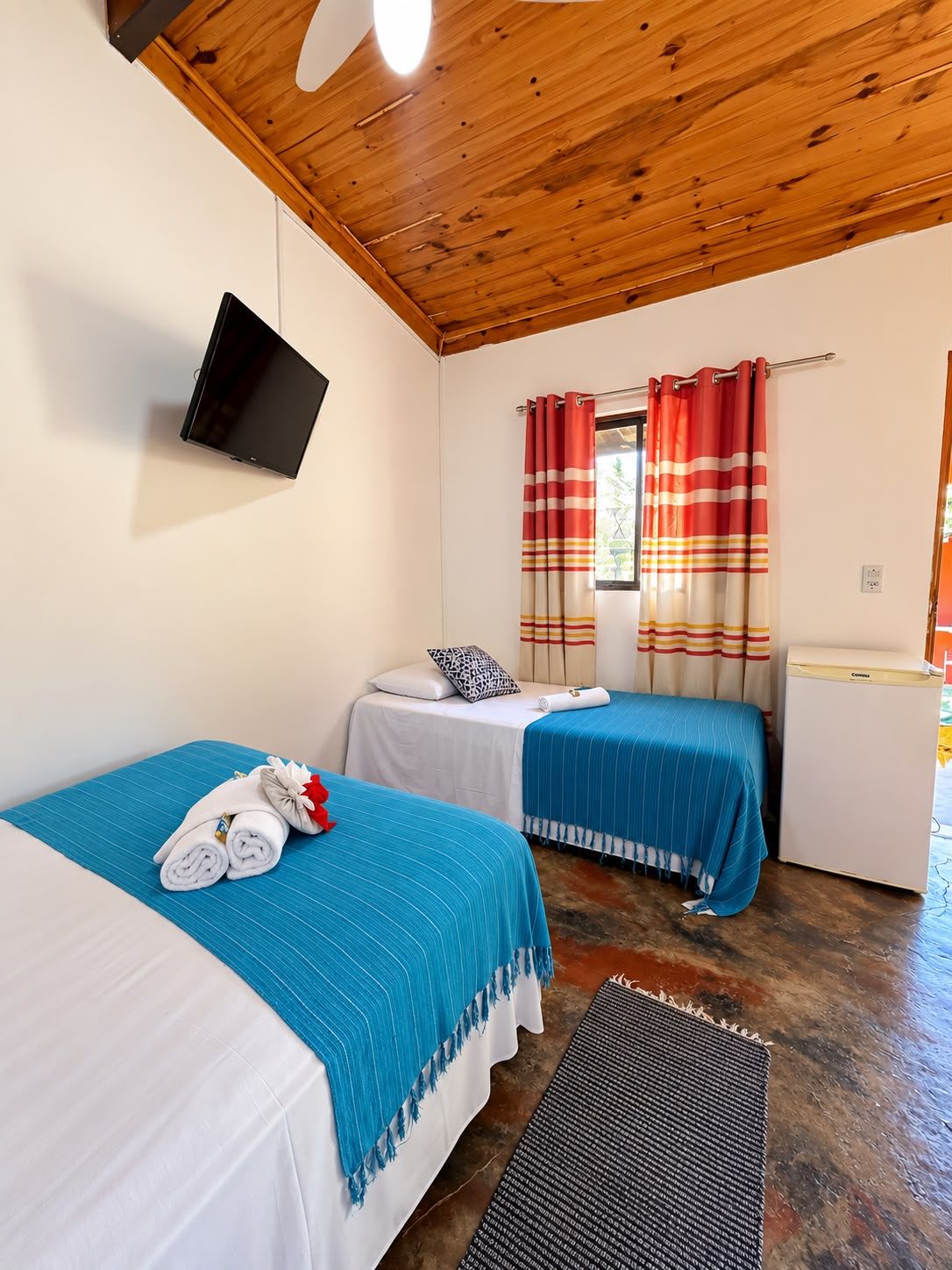 Habitación de hotel con dos camas, un televisor montado en la pared, una ventana con cortinas a rayas y un refrigerador pequeño.