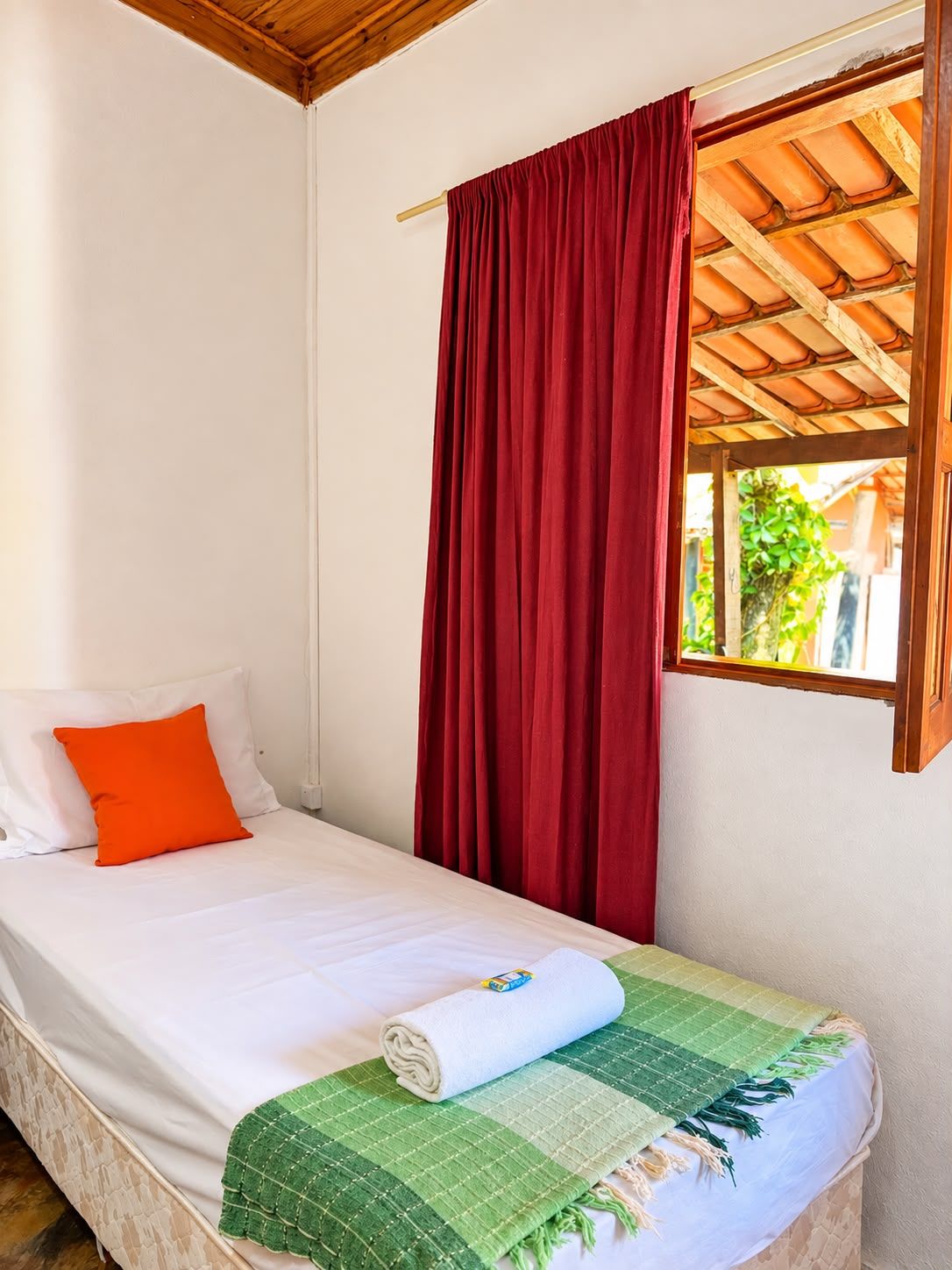 A single bed with white bedding and an orange pillow, a red curtain, and a window with a wooden ceiling.