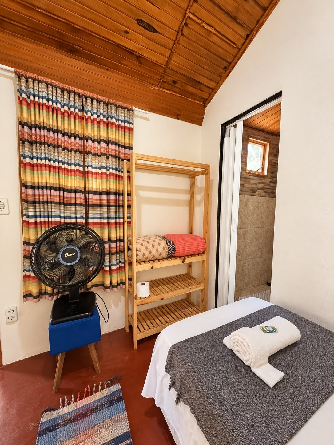 Un dormitorio con una cama nido, un ventilador de pie, una cortina estampada y una ventana pequeña.