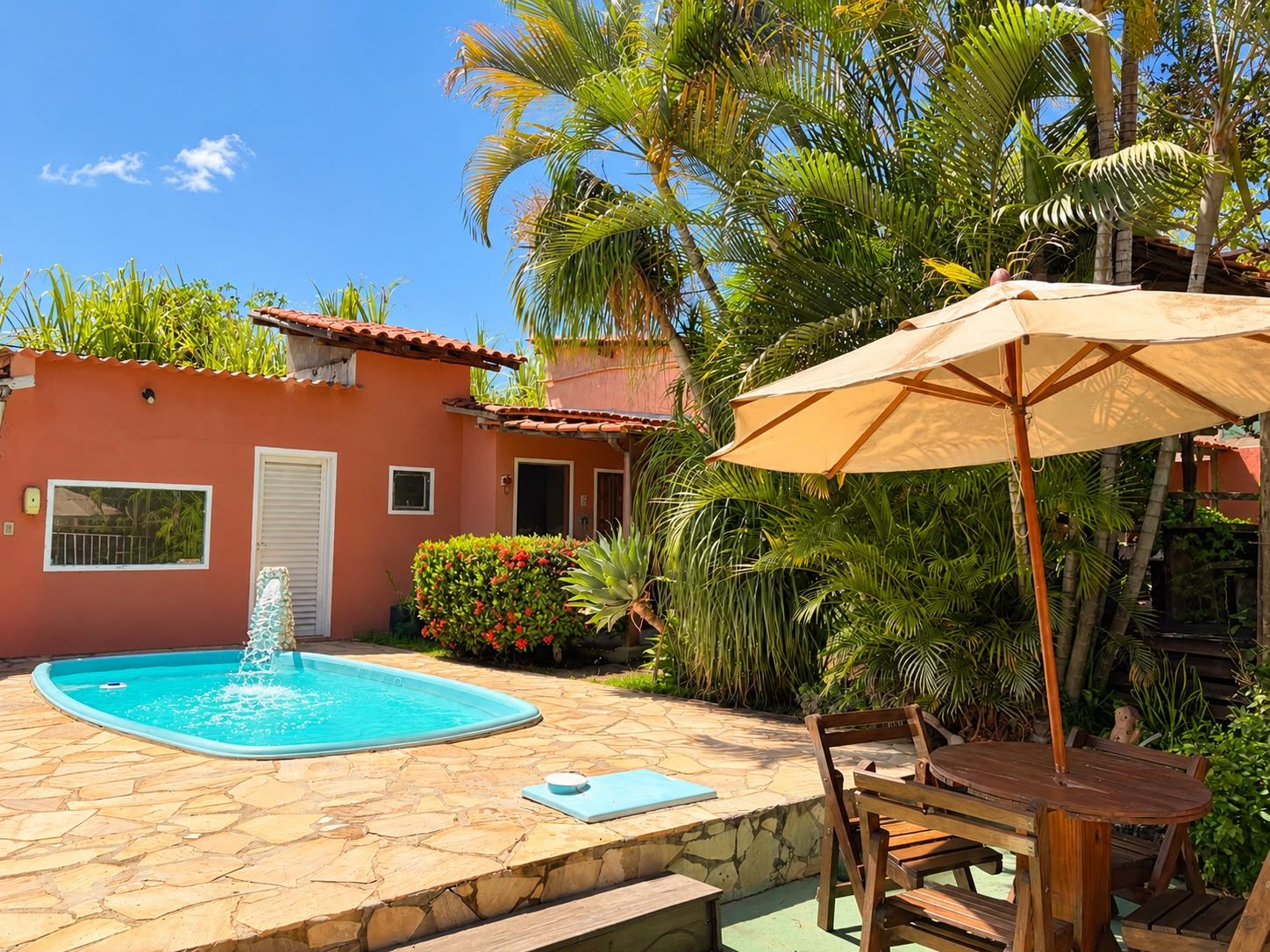 Uma área de piscina com um terraço de pedra, uma mesa de madeira com cadeiras e um guarda-sol.