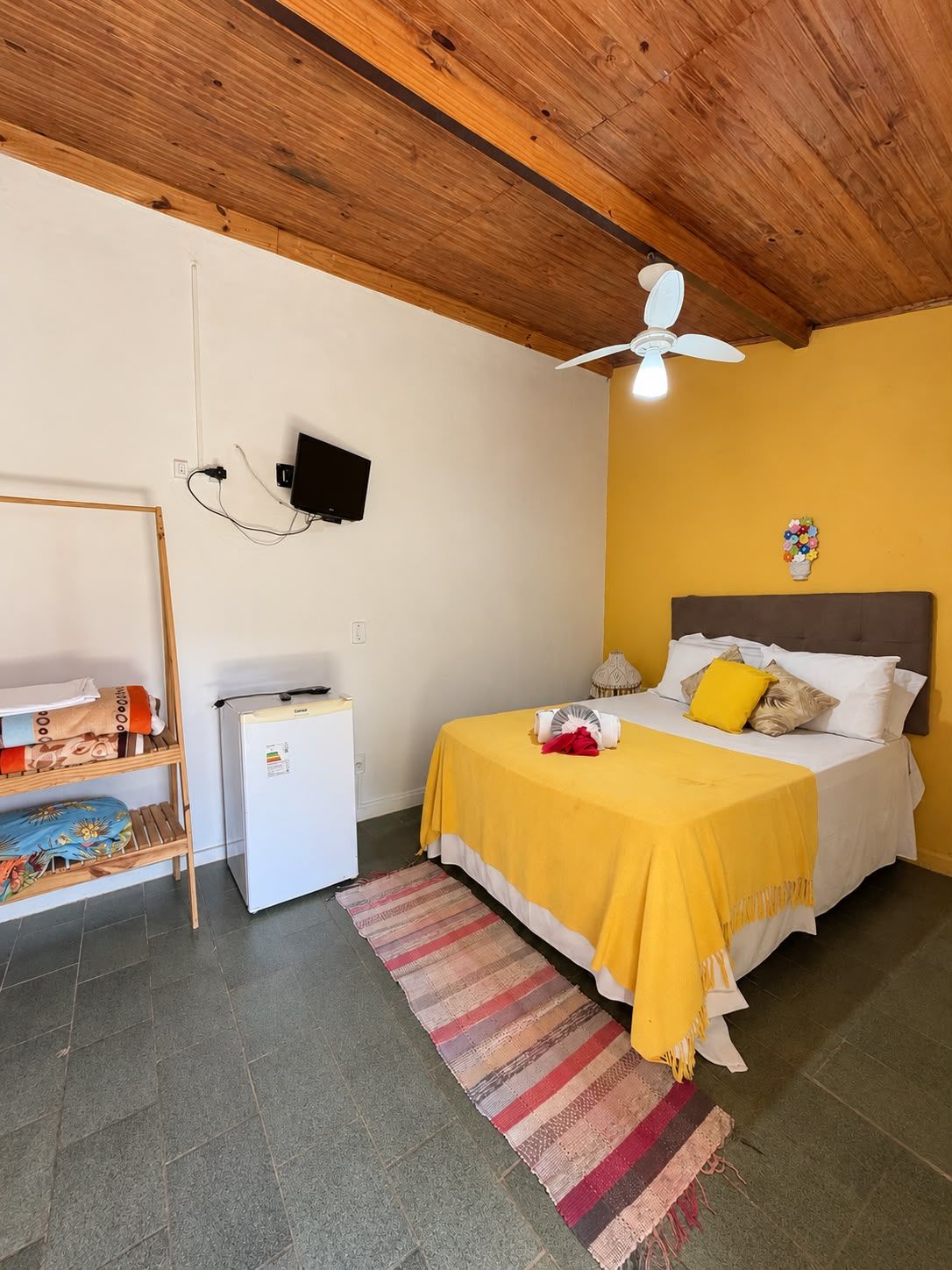 A bedroom with a bed, a small white refrigerator, a wooden shelf, a ceiling fan, and a wall-mounted TV.