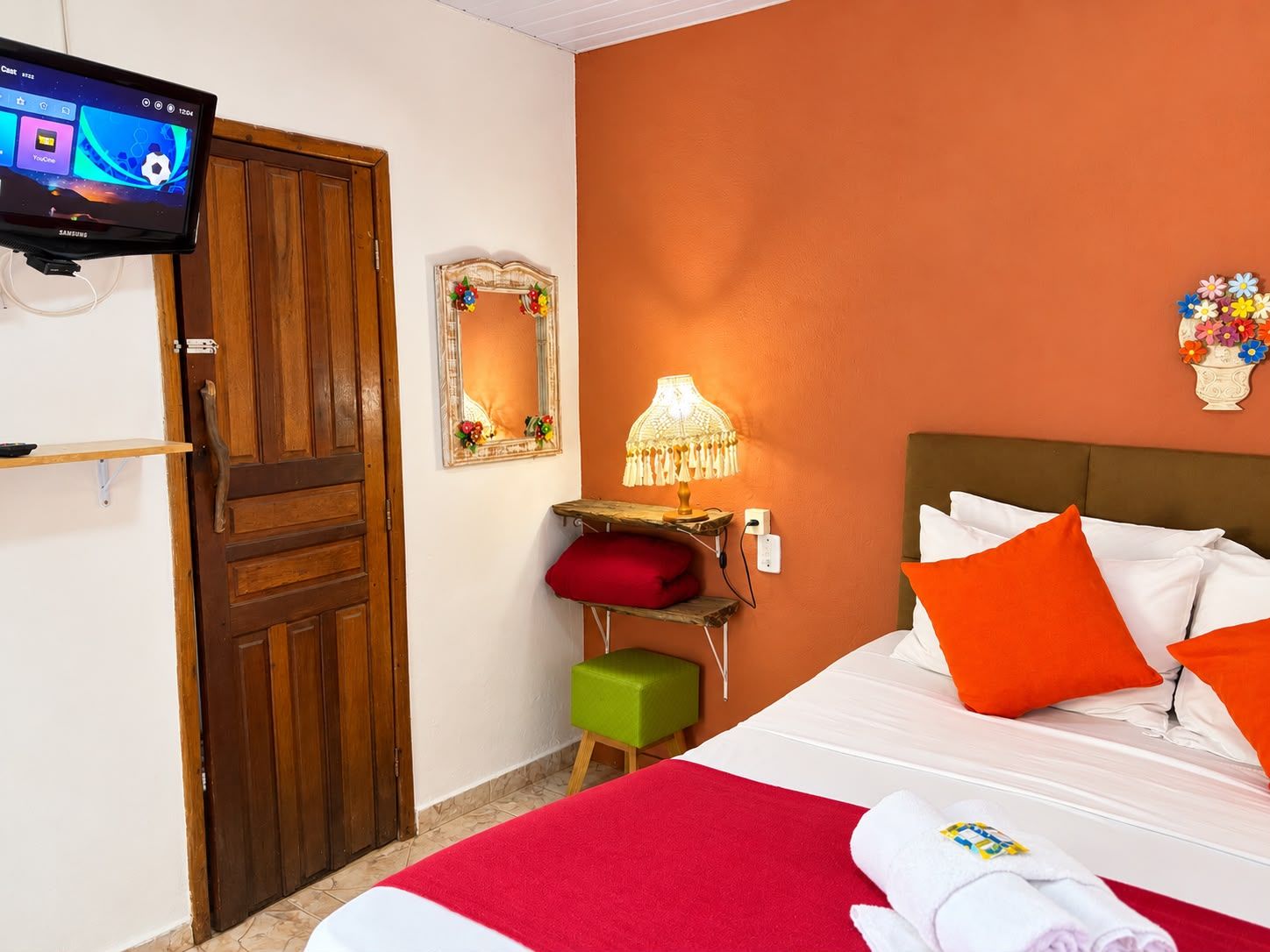 A hotel room with a bed, a TV mounted on the wall, a wooden door, a small table with a lamp, and a mirror with a floral decoration.
