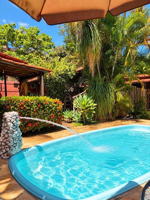 A swimming pool with a stone wall, surrounded by tropical plants and a wooden pergola.