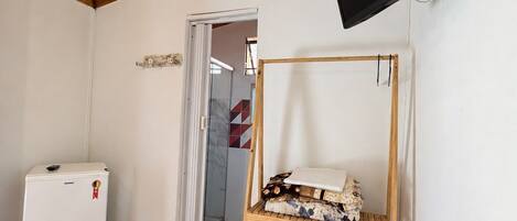 A room with a wooden ceiling, a television mounted on the wall, a small white refrigerator, a wooden shelf with items, and a tiled floor.