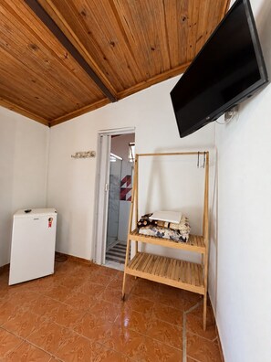 A room with a wooden ceiling, a television mounted on the wall, a small white refrigerator, a wooden shelf with items, and a tiled floor.