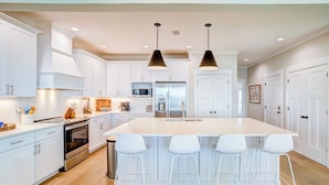 A modern kitchen with white cabinetry, a large island with bar stools, stainless steel appliances, and pendant lighting. - Lux 6BR, Lagoon Front, Private Heated Pool, Gulf Views, Boat Dock, Walk To Beach (Gulf Shores)