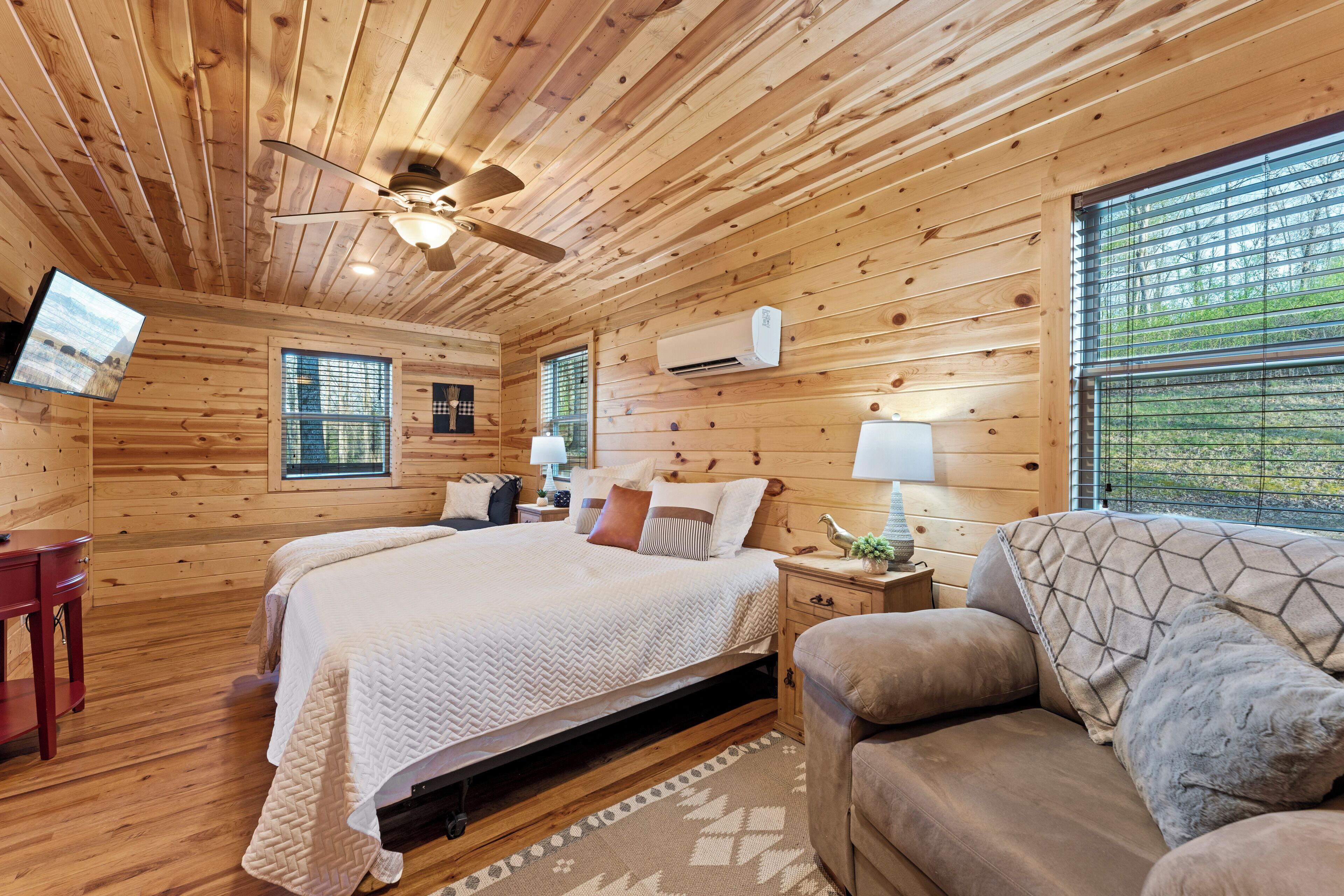 A wooden cabin bedroom with a bed, a sofa, a TV, and a ceiling fan.