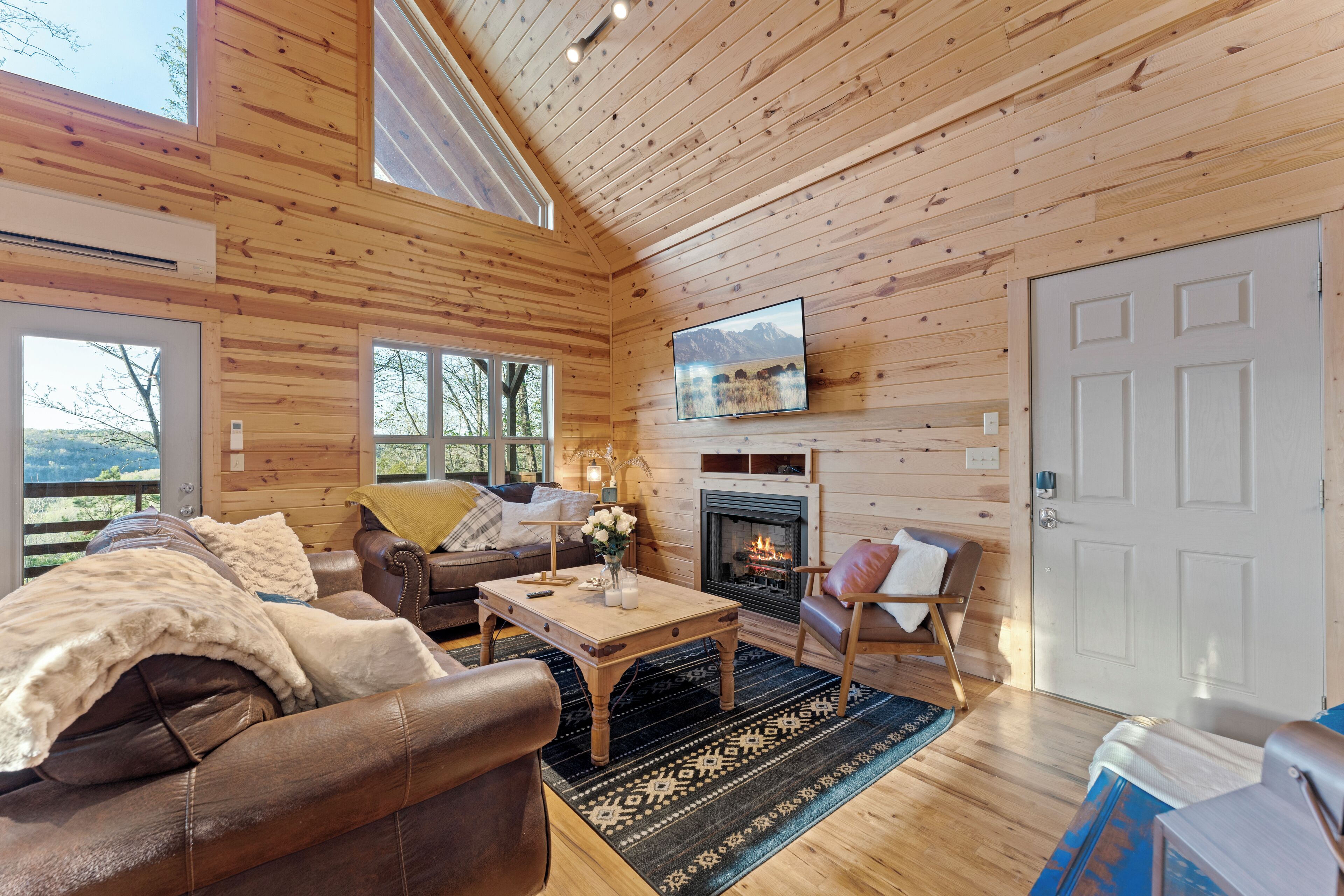 A cozy log cabin interior with a fireplace, wooden walls, a television, and a large window overlooking a scenic view.