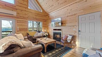 A cozy log cabin interior with a fireplace, wooden walls, a television, and a large window overlooking a scenic view.