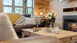 L’intérieur chaleureux d’un chalet, avec un décor en bois, une cheminée, un canapé agrémenté de coussins, une table basse ornée de bougies et d’un vase de fleurs, et un téléviseur fixé au mur.