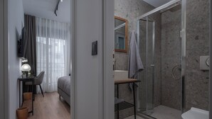 A modern hotel room with a bed, a desk with a lamp, a shower area with a glass door, and a bathroom with a toilet and sink.