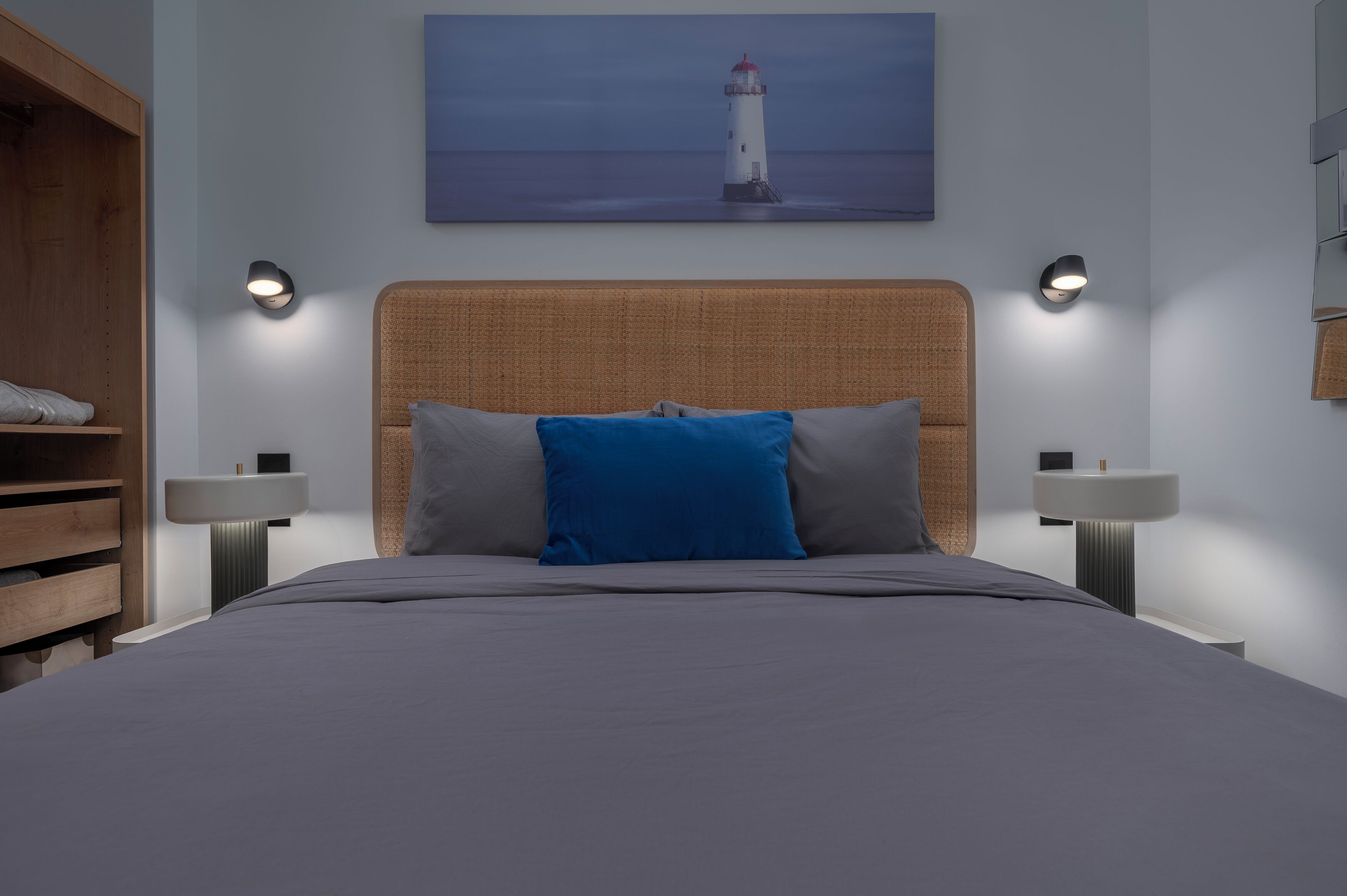 A modern bedroom with a large bed, two bedside tables, a wall-mounted painting of a lighthouse, and a wooden wardrobe.