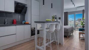A modern kitchen with white cabinets, a black countertop, and a bar area with white stools. There is a bed with a grey headboard in the adjacent room, and a balcony with a view of the city.