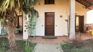 Una casa con puerta marrón, un porche pequeño y un jardín con plantas y un árbol.
