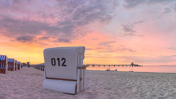 Een strandstoel met het nummer 012 op een zandstrand, met een pier op de achtergrond.