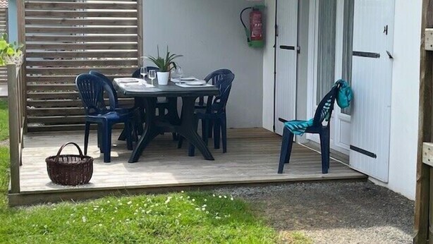 A covered patio area with a table set for four, surrounded by chairs, a small potted plant, and a basket. - 2 - Le Martin-Pêcheur - Village vacances (Maché)