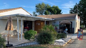 A small building with a white exterior and a tiled roof, featuring a covered outdoor seating area with chairs and a table, surrounded by greenery and a gravel path. - 2 - Le Martin-Pêcheur - Village vacances (Maché)