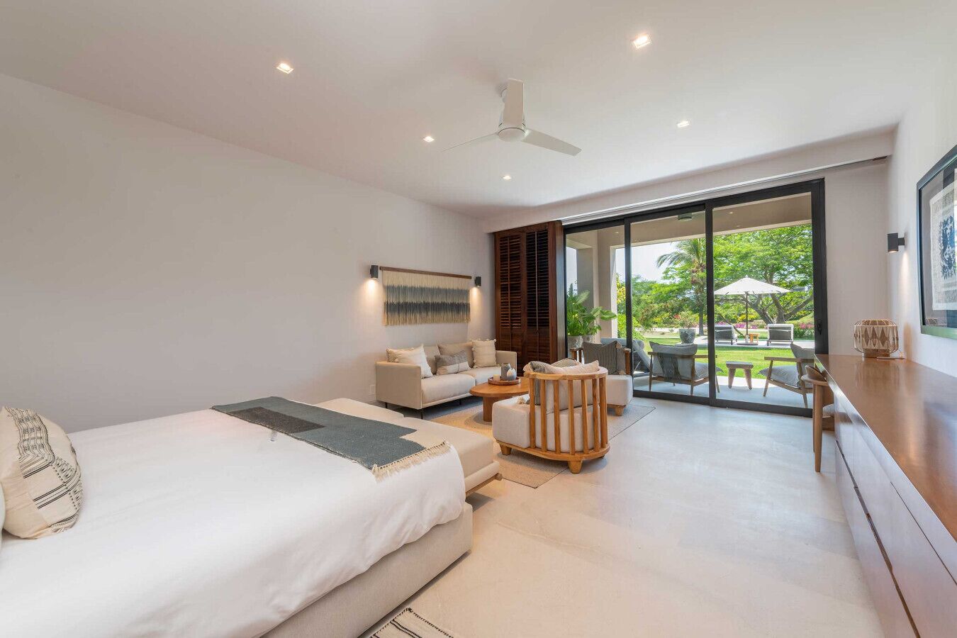 A modern bedroom with a large bed, a sofa, a wooden chair, and a dining area with a view of the garden.