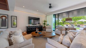 A modern living room with a large sofa set, a wooden coffee table, and a black entertainment unit. There is a large window with a view of a garden and outdoor seating area.